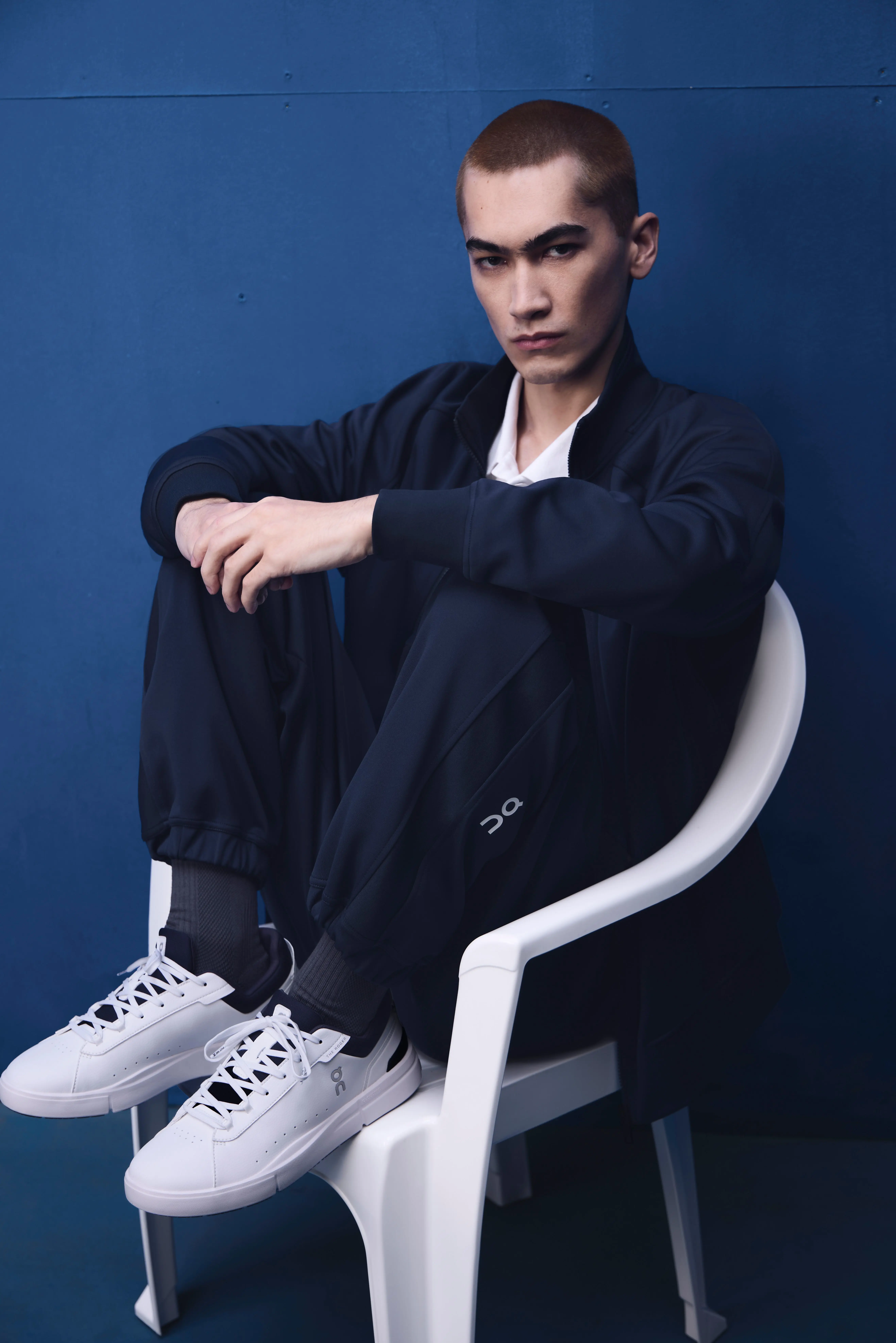 A person sitting on a white plastic lawn chair, wearing On gear, feet up on the chair.