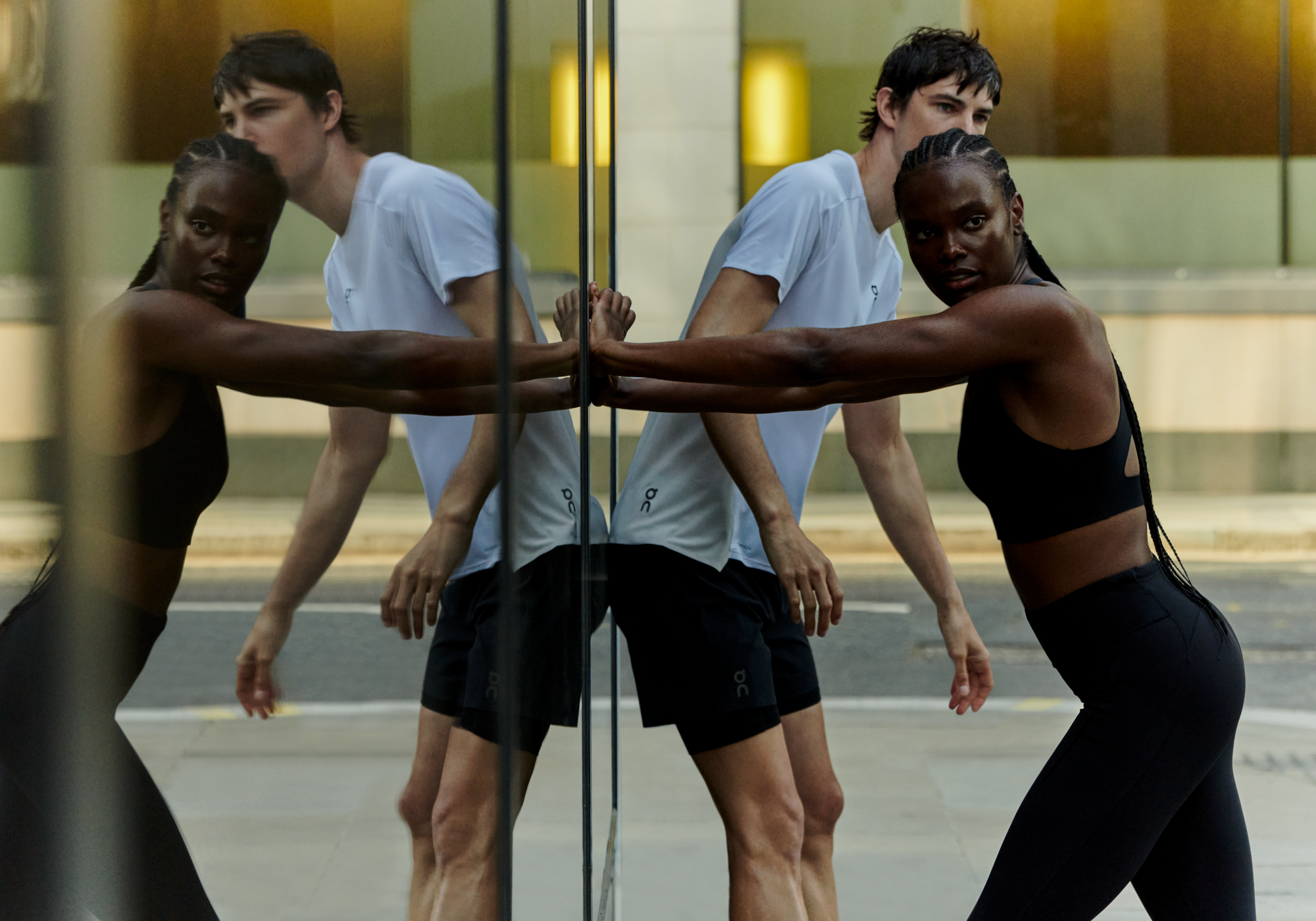 Two athletes leaning on a reflective glass surface outdoors while taking a break from a run, wearing On performance apparel.