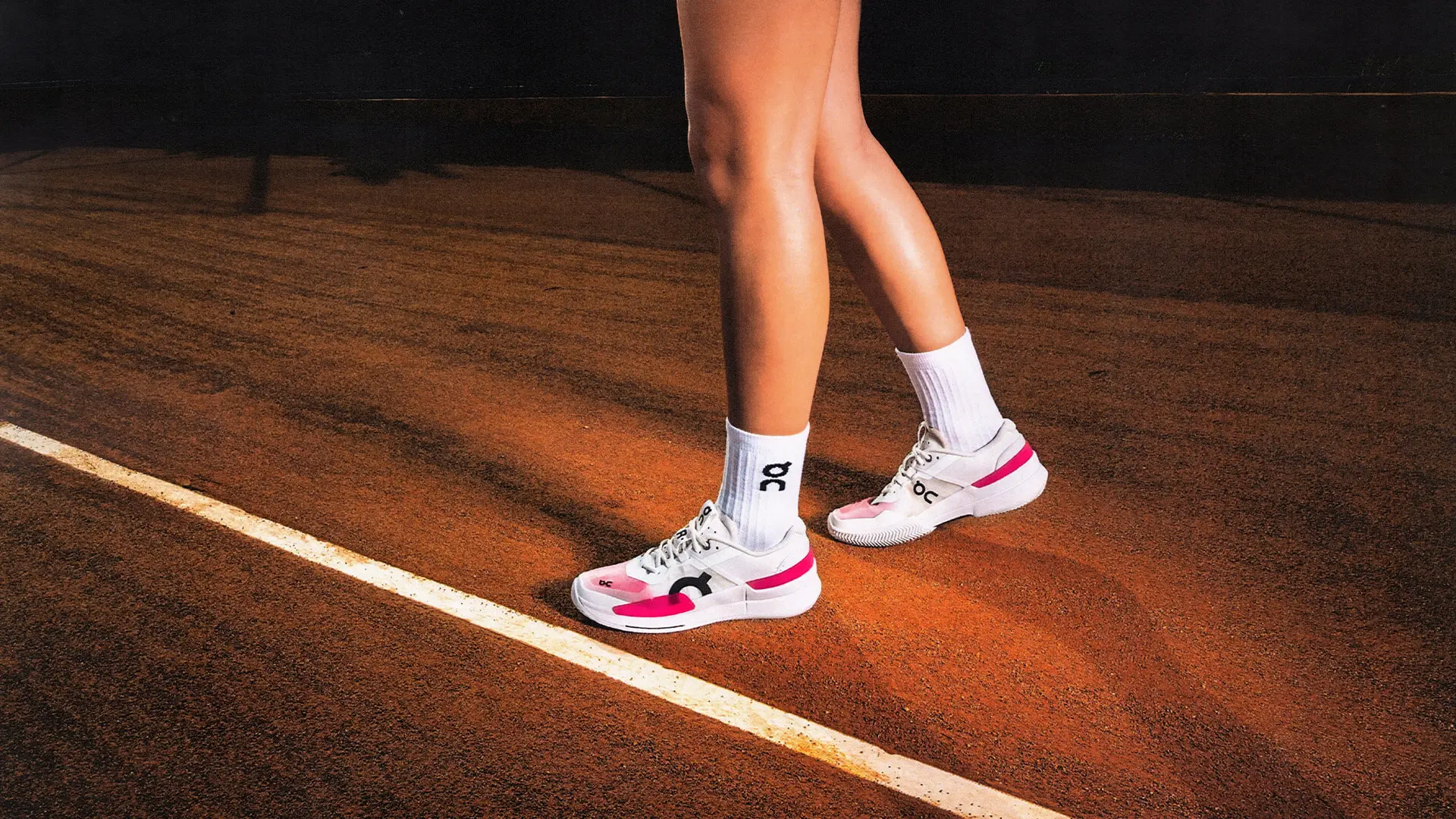 A woman wearing On clay tennis shoes stands on a clay court.