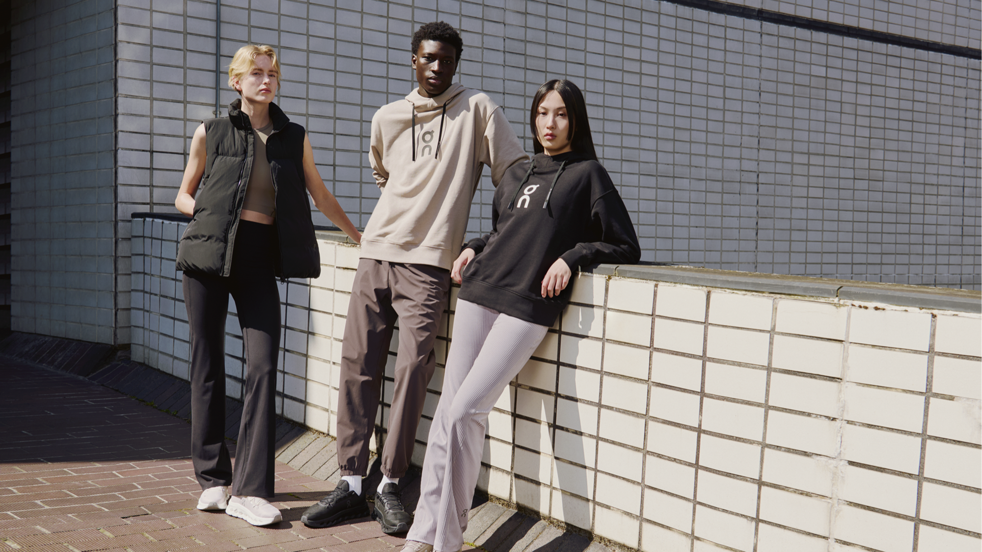 Three people wearing On gear while leaning against a brick wall outside and looking seriously at the camera.