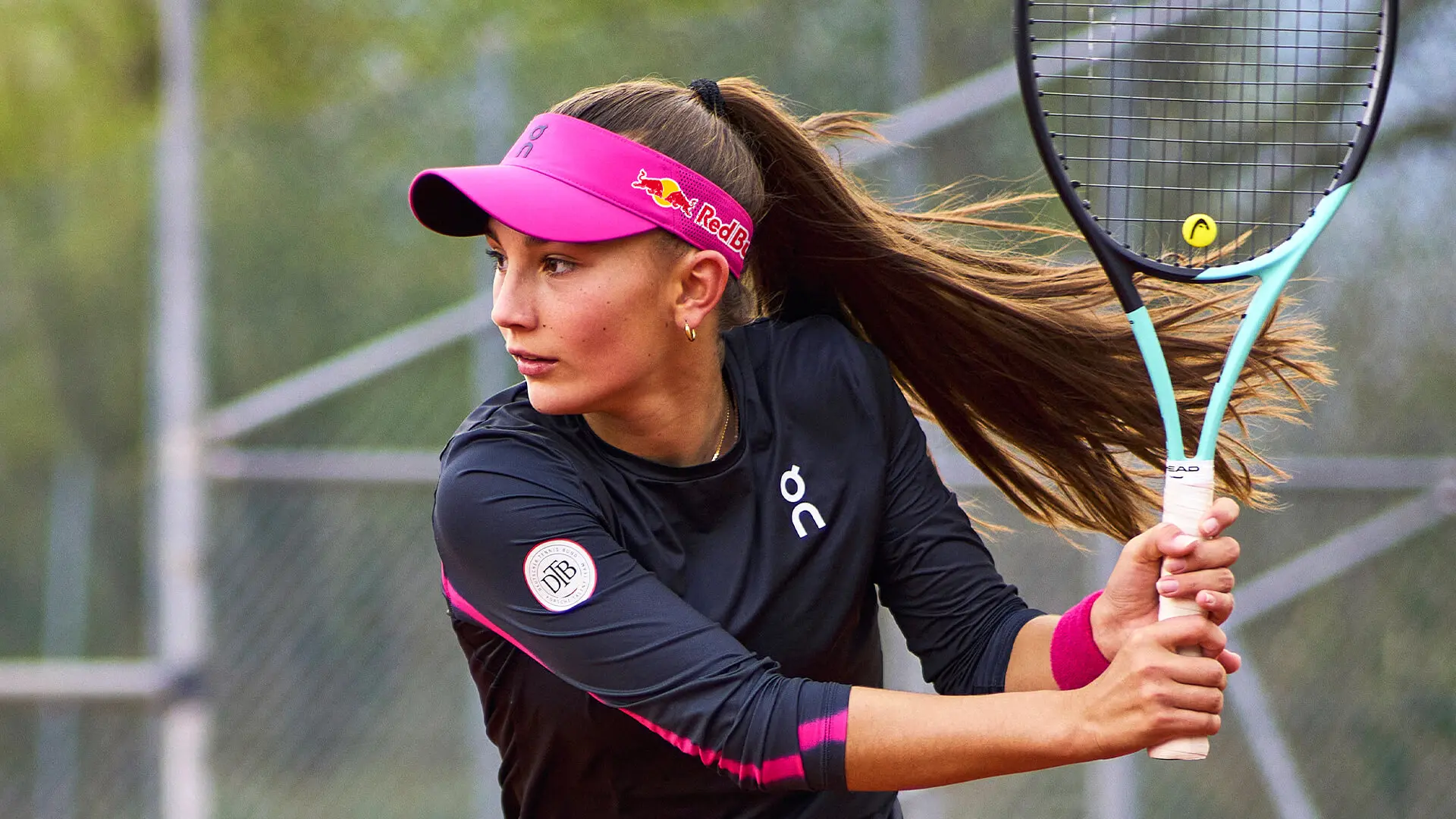 Julia Stusek in a pink visor prepares to return the tennis ball. 
