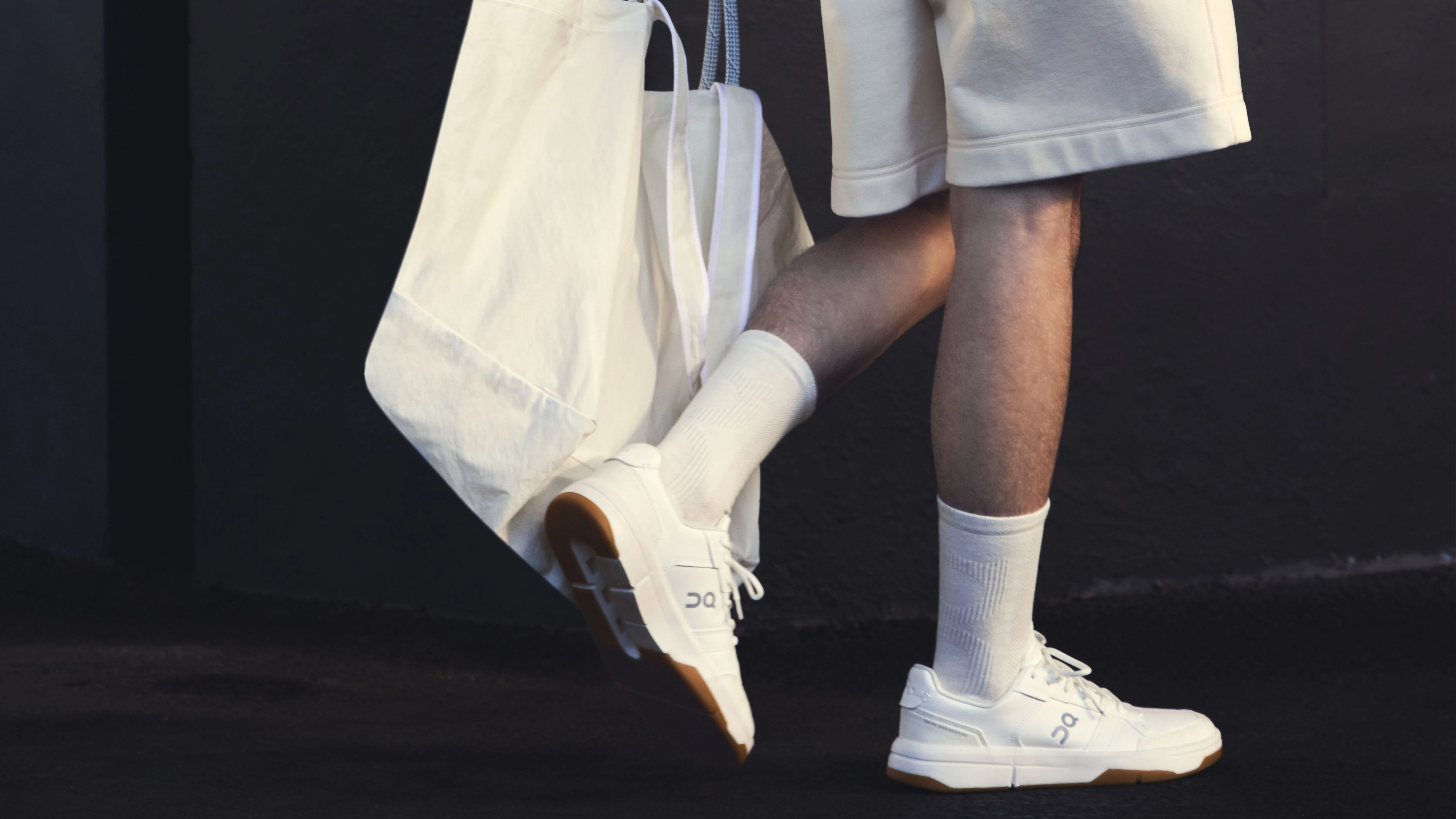 A person pictured from the hip down, wearing casual shorts, socks and On sneakers, carrying a tote.