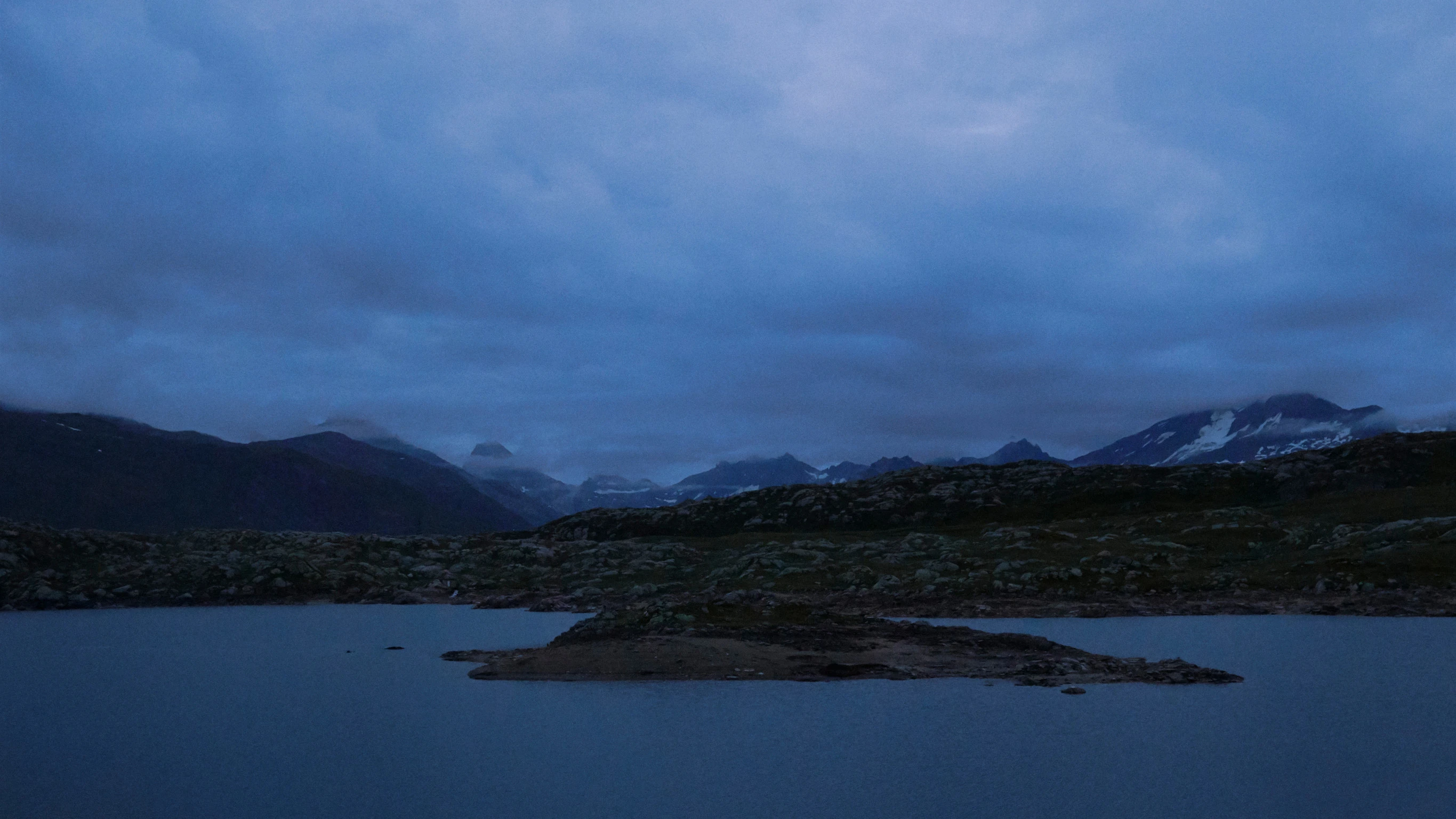 A scenic view, across a lake toward dark hills on a cold day, with snow-tipped mountains in the distance.