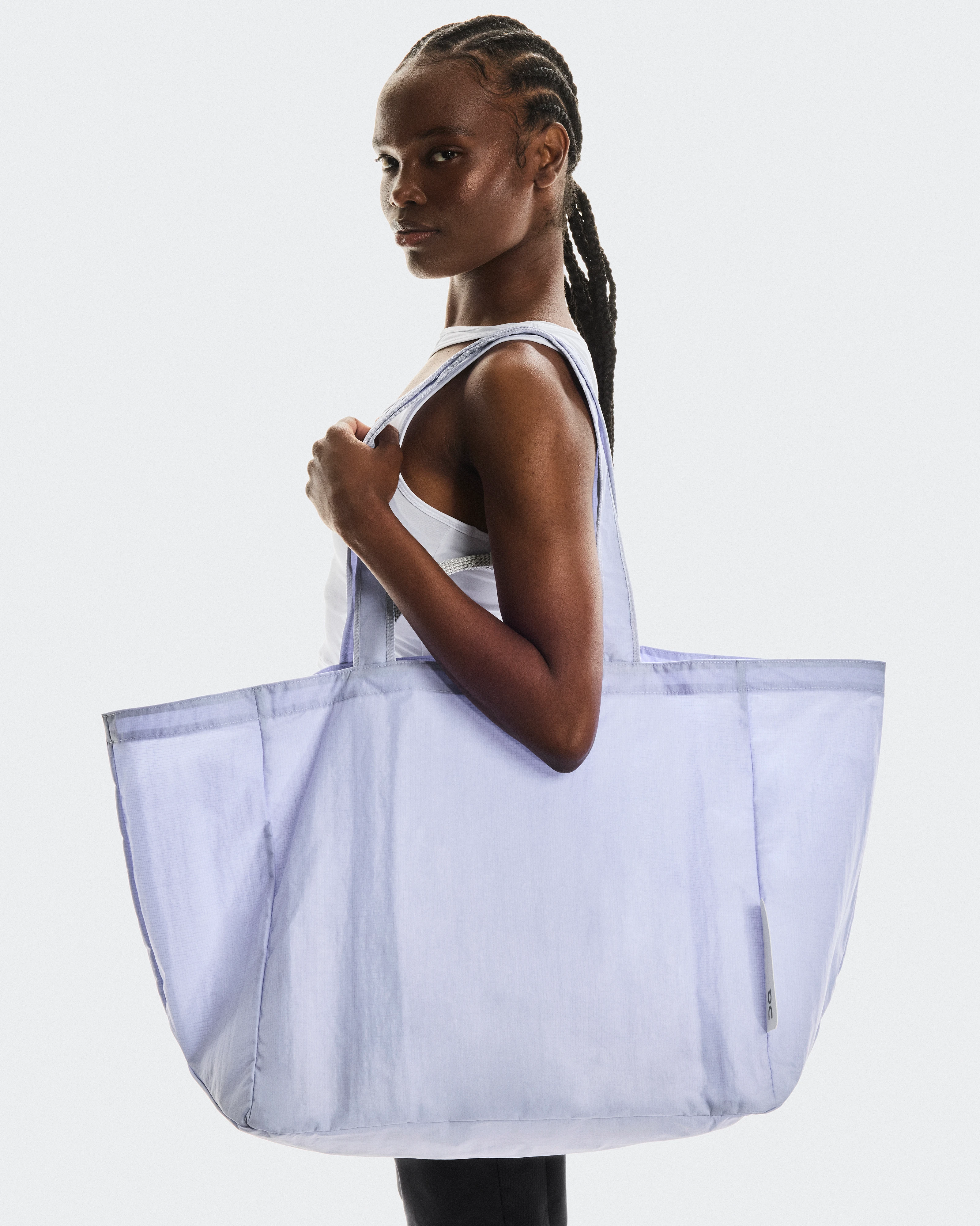 A woman with braided hair in a white tank top holds a large purple On mesh tote bag against a light gray background.