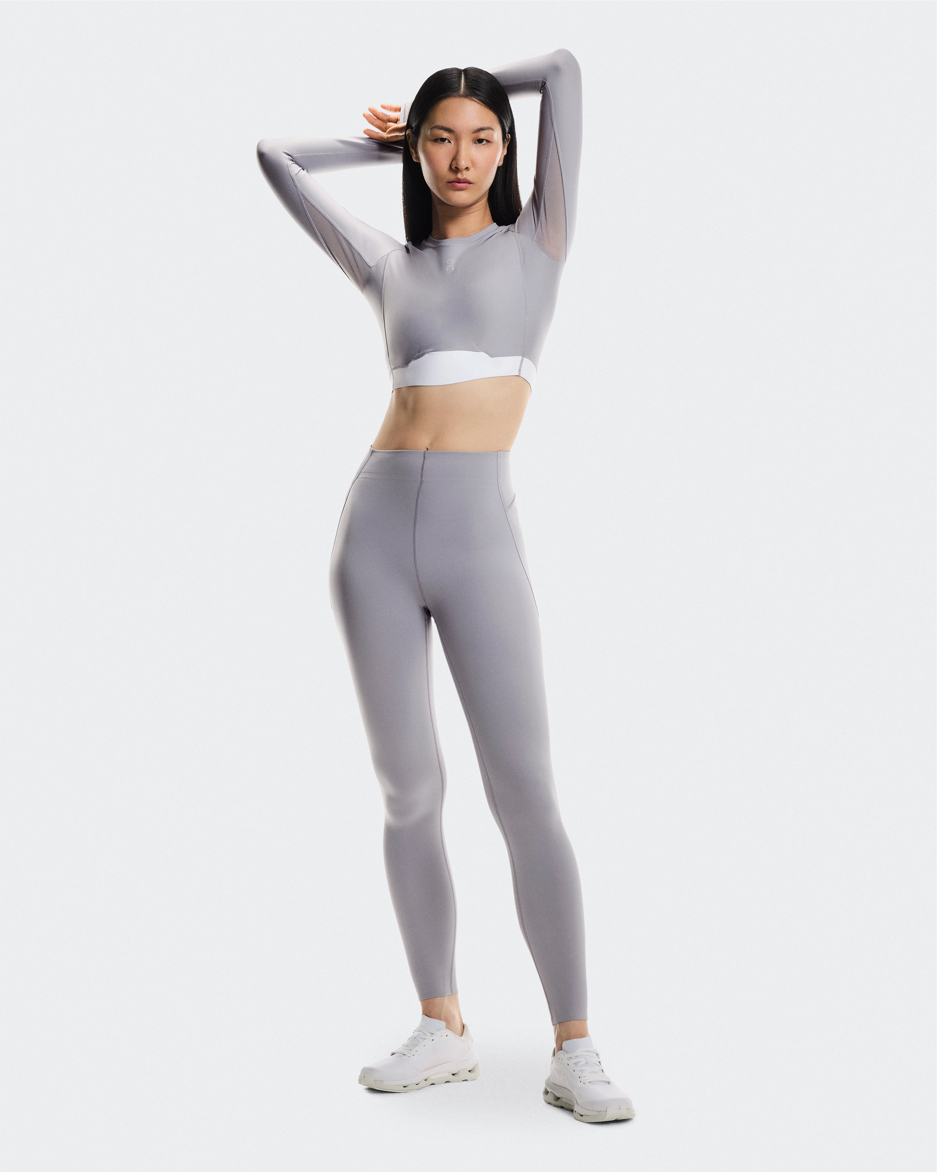 Woman wearing a tight gray long-sleeve, matching leggings, and On training shoes, standing while stretching with her arms behind her head.