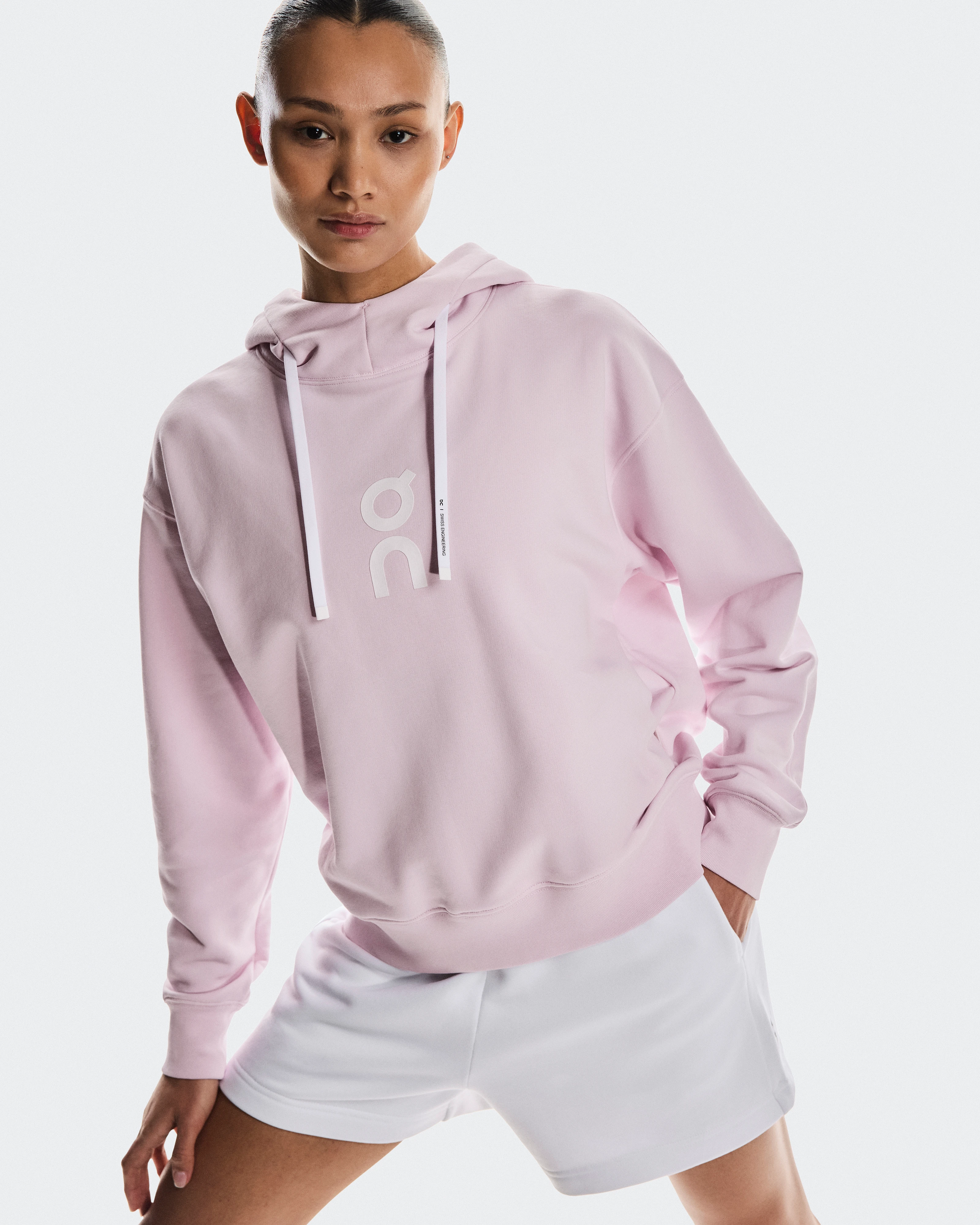 A woman wearing a pink On hoodie sweatshirt and white shorts, with a hand in her pocket.