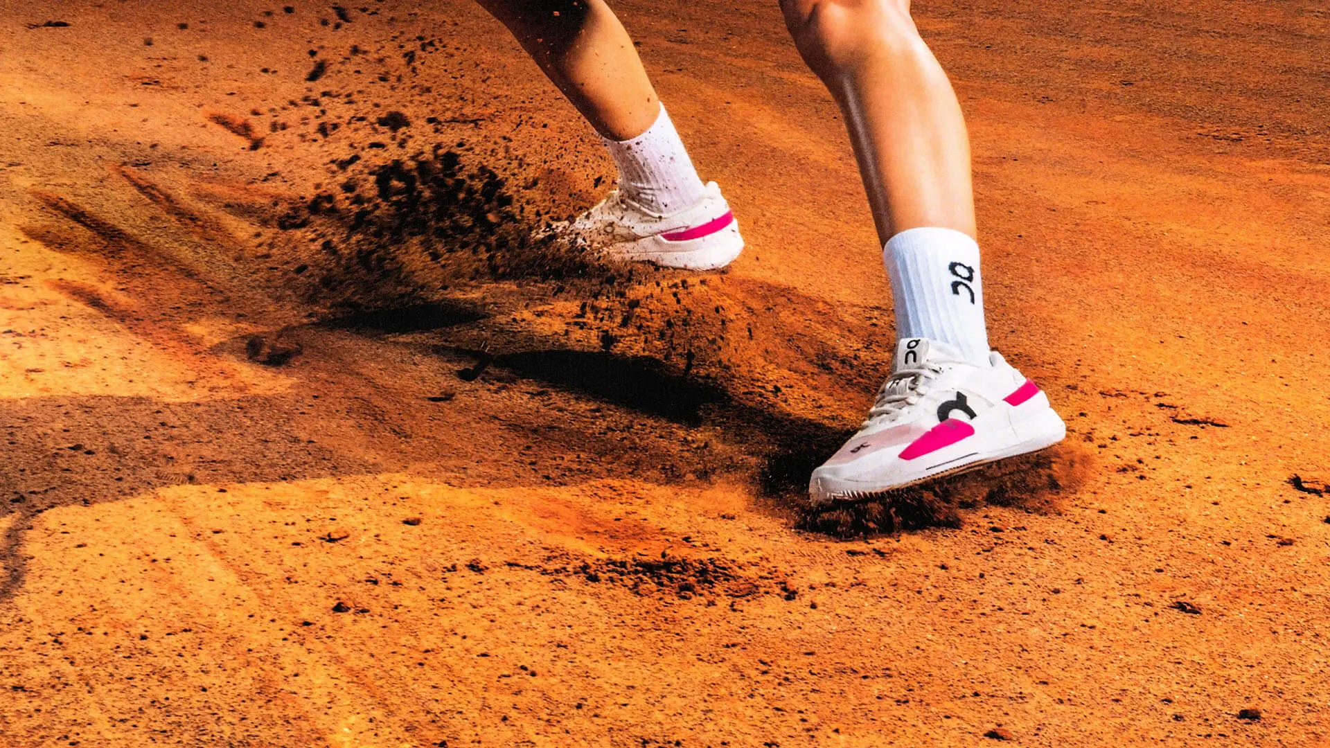 A person wearing On tennis shoes slides across a clay tennis court.