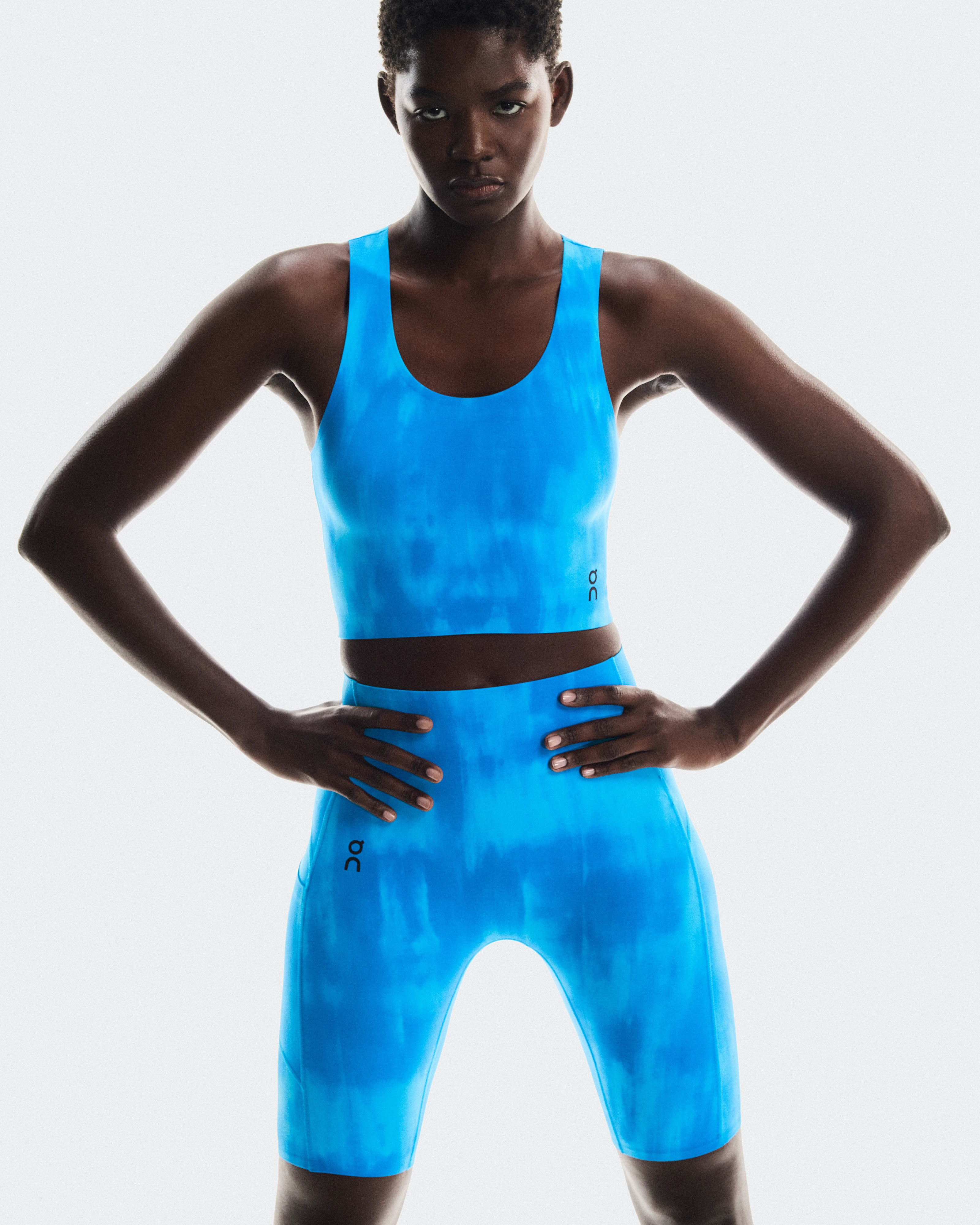 Woman wearing a bright blue On Running sports bra and matching bike shorts, standing with hands on hips.