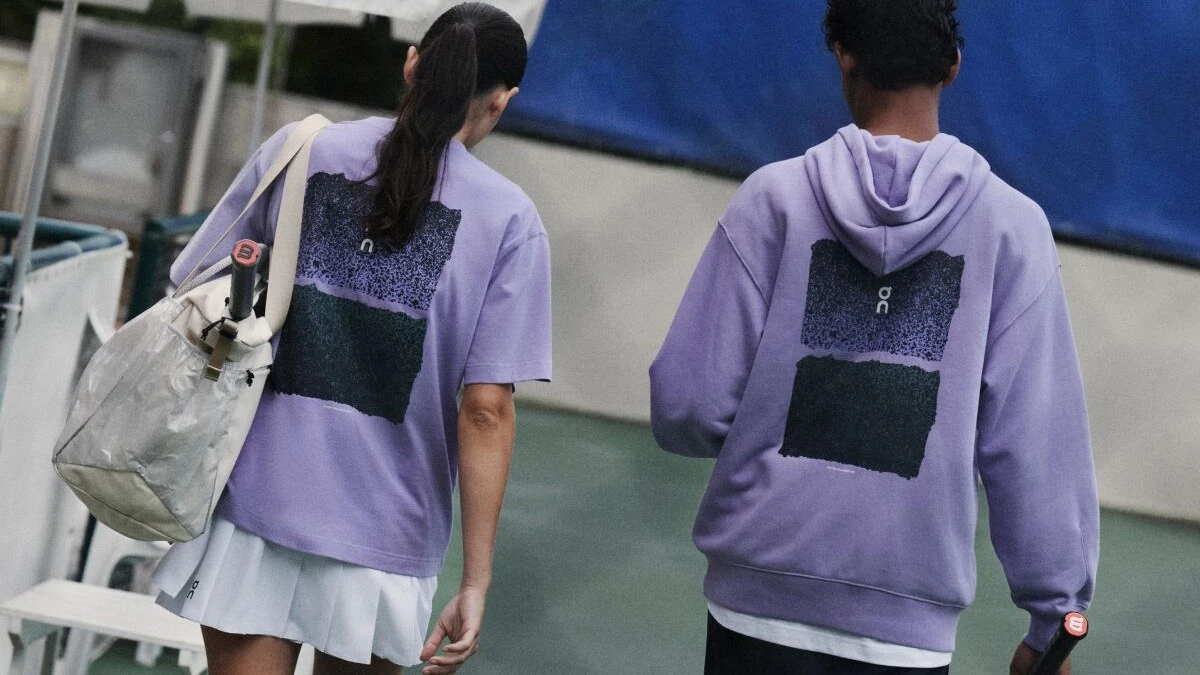 Two tennis players walking away courtside, both in purple On tops, a t-shirt and a sweatshirt.