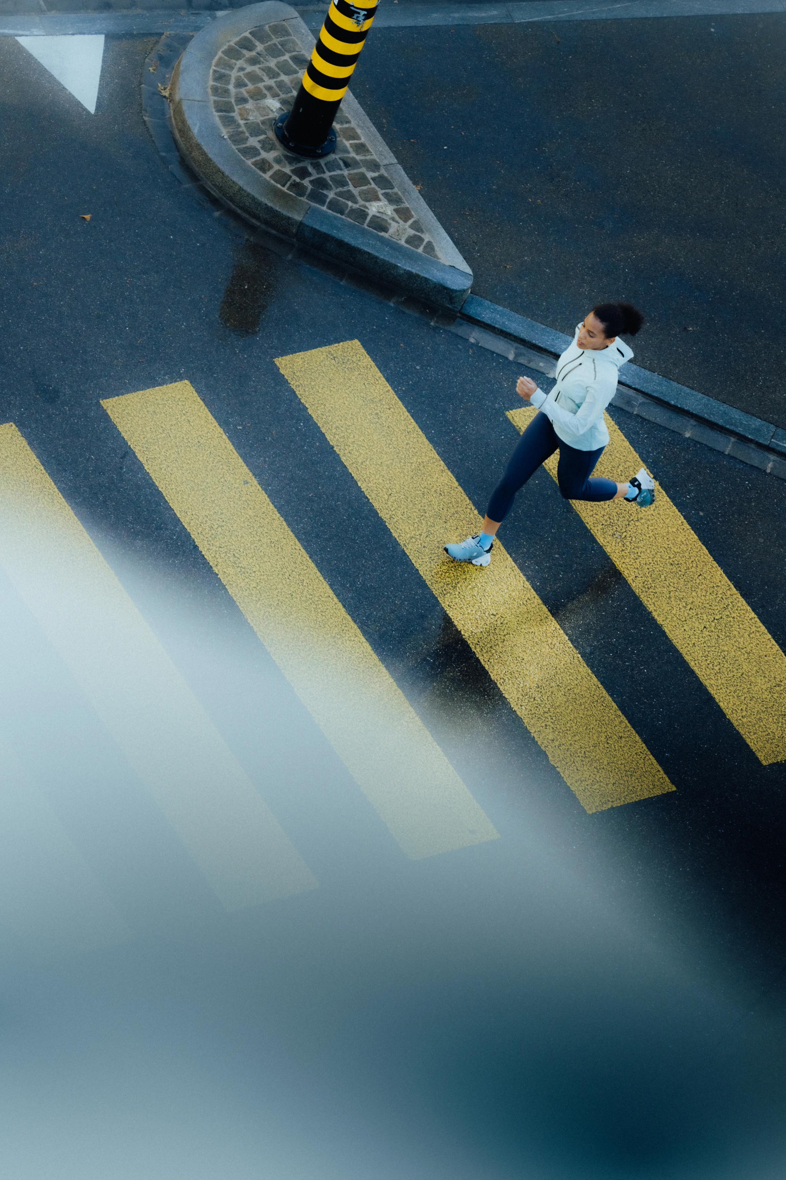 Overhead shot of a woman wearing water resistant On apparel, while running across a zebra crossing.