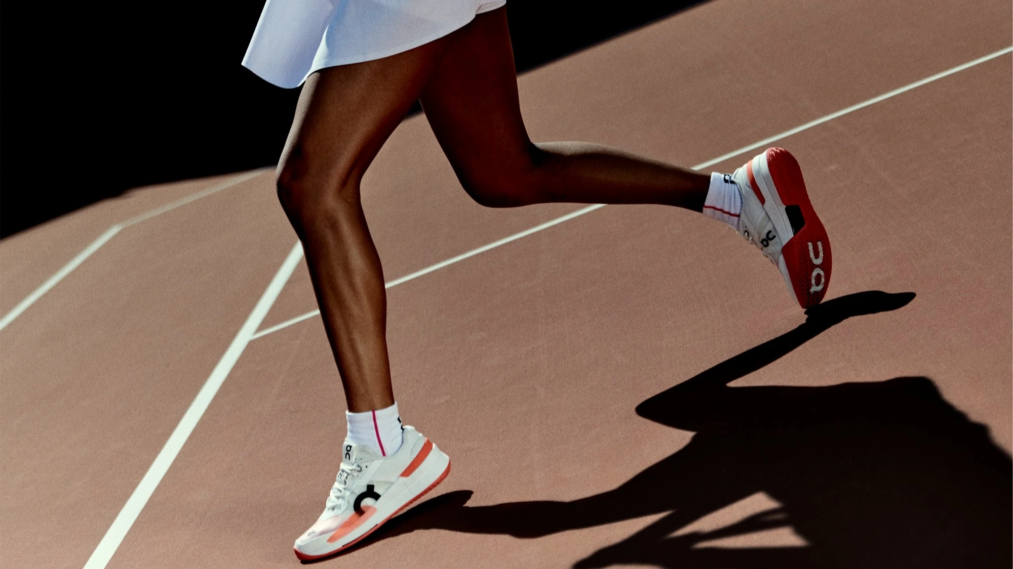 A woman in a white tennis skirt runs across a brown tennis court.