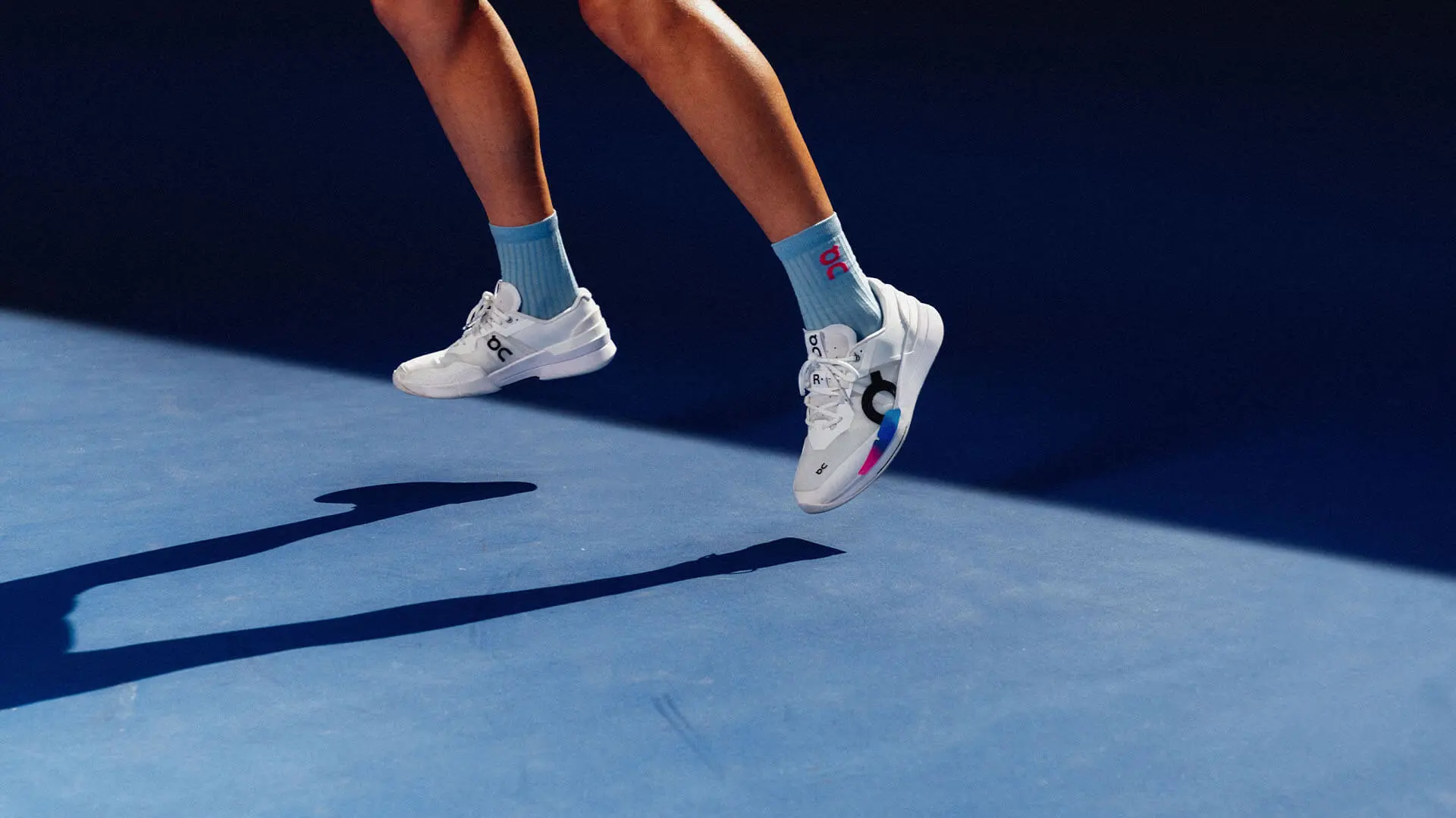 A person wearing On tennis shoes jumps off a blue hard tennis court.