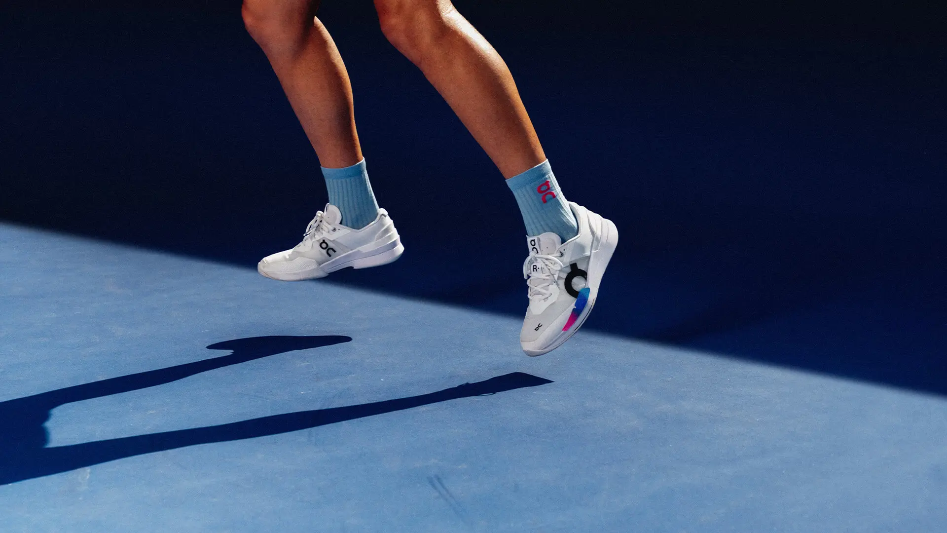 A person wearing On hard court tennis shoes jumps on a blue court.